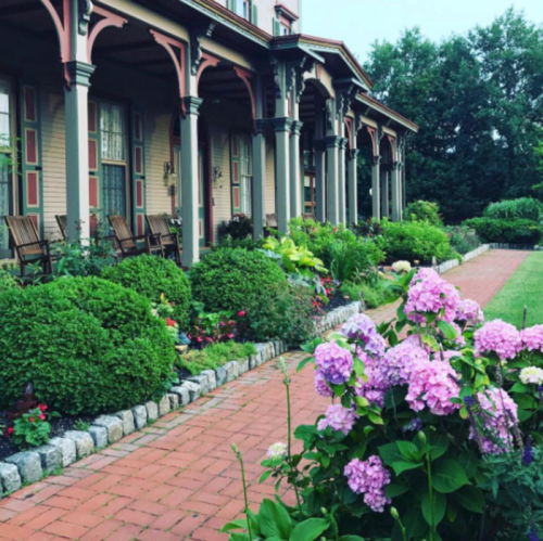 A charming house with a porch, surrounded by lush greenery and colorful flowers along a brick pathway.