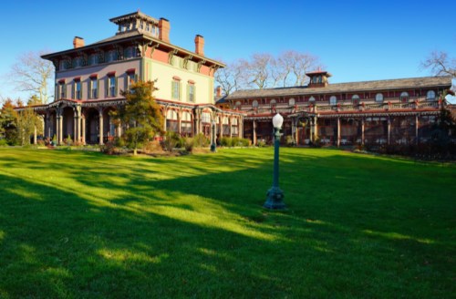 Victorian-style mansion with a large lawn, surrounded by trees and a clear blue sky.