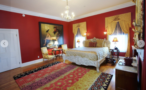 Luxurious bedroom with red walls, ornate bed, elegant decor, and a large painting. Warm lighting enhances the ambiance.