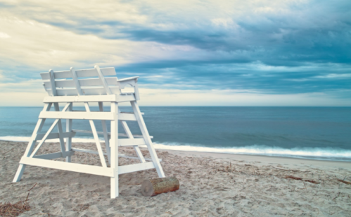 A white lifeguard chair on a sandy beach, overlooking a calm ocean under a cloudy sky.