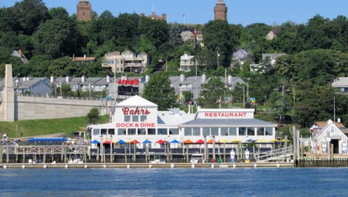 A waterfront restaurant with colorful umbrellas, surrounded by greenery and houses on a hillside.