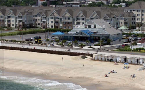 A beach scene with a restaurant, nearby buildings, and people relaxing on the sand under a clear sky.