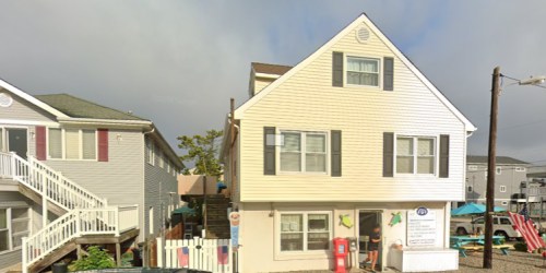 A two-story beach house with a light-colored exterior, surrounded by other buildings and outdoor seating areas.