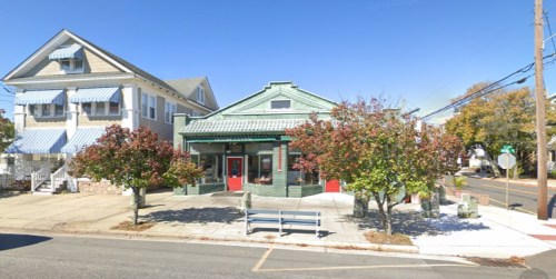 A green building with red doors, flanked by trees, next to a light-colored house with blue awnings on a sunny day.