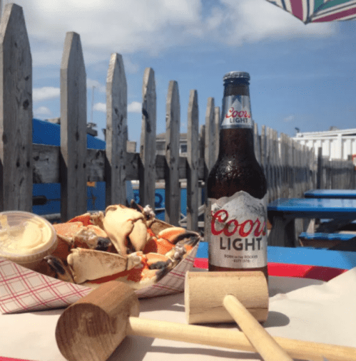 A bottle of Coors Light next to a crab dish and wooden mallets on a table with a blue sky and wooden fence in the background.