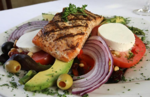 Grilled salmon atop a fresh salad with tomatoes, avocado, olives, red onion, and mozzarella, garnished with herbs.