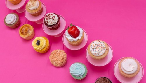 A colorful array of cupcakes on pink stands, featuring various toppings and flavors, arranged in a line.