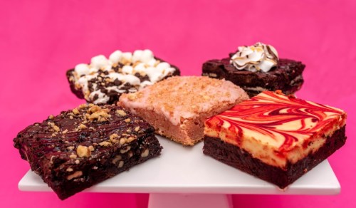A variety of colorful brownies on a white plate, including chocolate, strawberry, and swirled flavors, against a pink background.