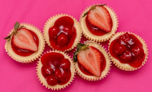 Six mini cheesecakes topped with strawberries and cherry gelatin on a pink background.