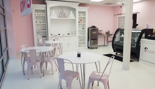 A pastel pink dessert shop interior with white tables, chairs, and a display case filled with treats.