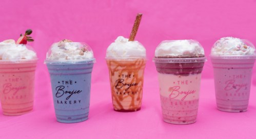 Five colorful milkshakes in clear cups with whipped cream, topped with various garnishes, against a pink background.