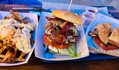 A tray with three food items: a large burger with bacon, lettuce, and tomato, loaded fries, and a sandwich.