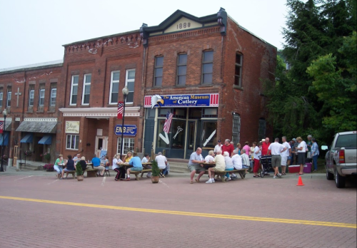 A group of people gathered outside the American Museum of Cutlery, with tables set up on a brick street.