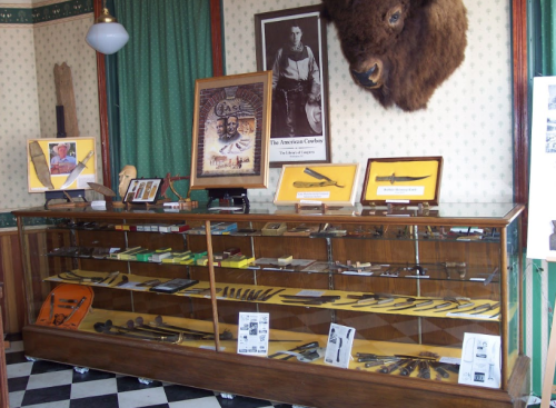 Display case filled with various knives and tools, featuring a mounted animal head and cowboy-themed decor in the background.