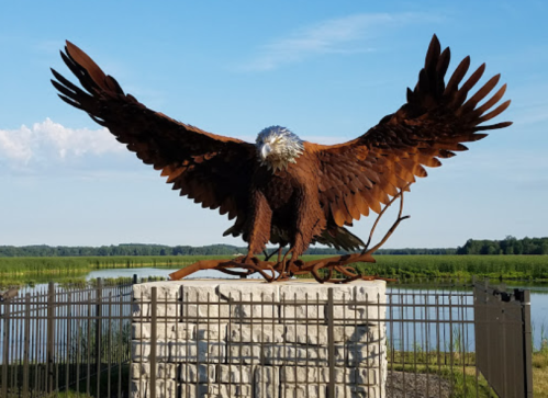 A large metal sculpture of an eagle with outstretched wings perched on a stone base, set against a scenic landscape.