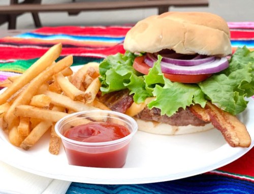 A juicy burger with lettuce, tomato, onion, and bacon, served with fries and ketchup on a colorful tablecloth.