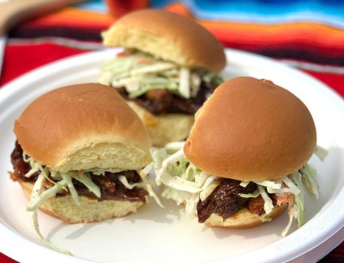 Three pulled pork sliders topped with coleslaw on a white plate, set against a colorful woven background.