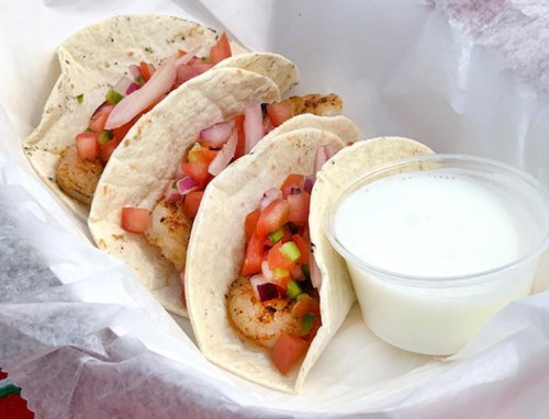 Three shrimp tacos in soft tortillas, topped with diced tomatoes and onions, served with a small cup of white sauce.