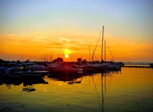 Sunset over a marina with boats silhouetted against the vibrant orange and yellow sky reflecting on the water.