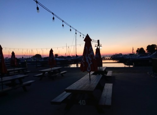 Silhouette of picnic tables with umbrellas at a marina during sunset, with boats and string lights in the background.