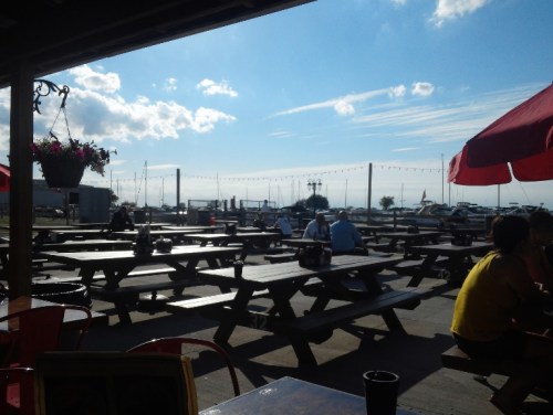Outdoor dining area with picnic tables, overlooking a marina and blue sky with clouds. People are seated, enjoying the view.