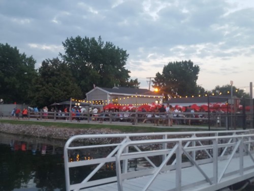 A lively outdoor dining area by a waterway, with string lights and red umbrellas, filled with people enjoying the evening.