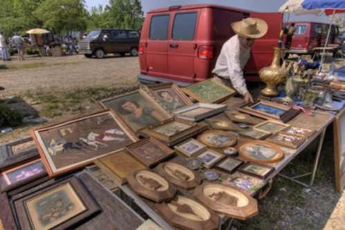 A vendor arranges framed artwork and antiques on a table at an outdoor market, with a red van in the background.