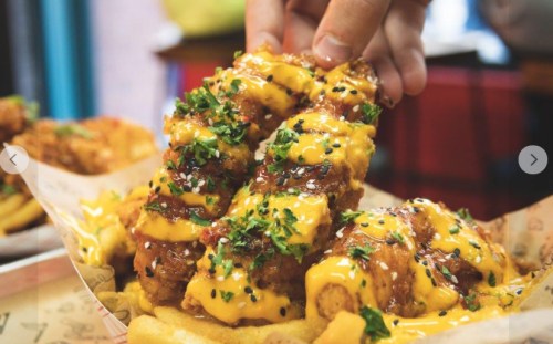 A hand holds a crispy chicken piece topped with cheese and herbs, served on a waffle with a side of fries.