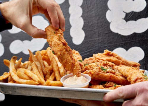 A hand reaches for a crispy chicken tender above a tray of golden fries and dipping sauce.