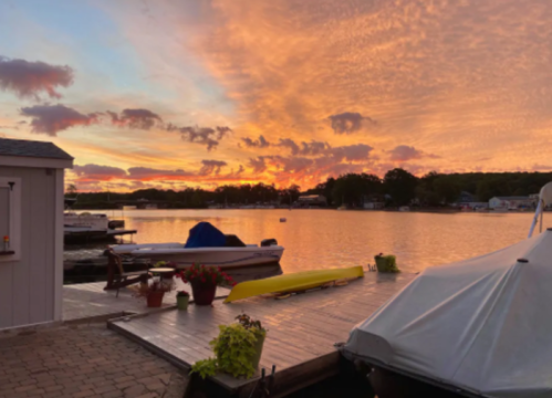 A serene lakeside scene at sunset, featuring colorful clouds, boats, and a wooden dock with potted plants.