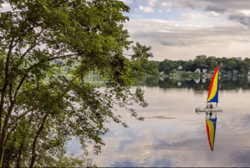 A colorful sailboat glides across a calm lake, surrounded by trees and reflecting the sky and shoreline.