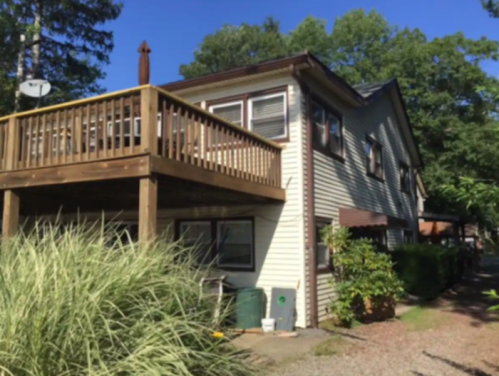 A two-story house with a wooden deck, surrounded by greenery and a gravel path on a sunny day.