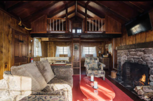 Cozy cabin interior with wooden walls, a stone fireplace, and comfortable seating, featuring a lofted area above.