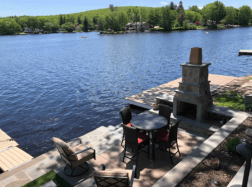 A serene lakeside view featuring a patio with a table, chairs, and a stone fireplace, surrounded by greenery.