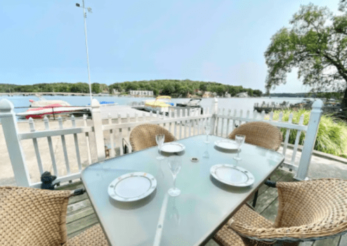 A table set for four on a deck overlooking a lake with boats and greenery in the background.
