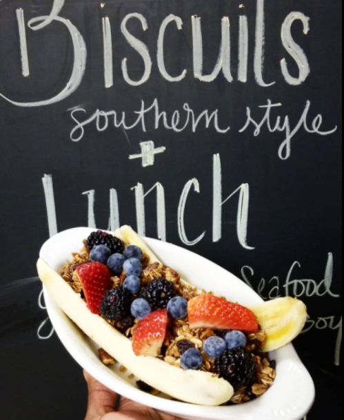 A hand holding a white dish filled with granola, berries, and banana slices, with a chalkboard menu in the background.