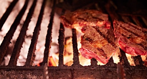 Grilled steaks sizzling on a barbecue grill, with flames and smoke in the background.