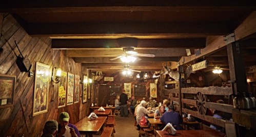 Cozy rustic restaurant interior with wooden beams, tables, and vintage posters, filled with diners enjoying their meals.