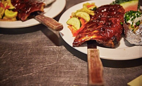 Two plates of barbecue ribs with sauce, served with pickles, salad, and a baked potato on a metal table.