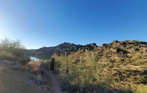 A panoramic view of a desert landscape with rocky hills and a calm lake under a clear blue sky.