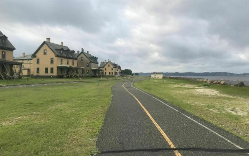 A winding path beside grassy areas and historic buildings under a cloudy sky near a body of water.