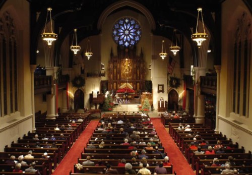 A view of a church interior filled with people, featuring a large stained glass window and festive decorations.