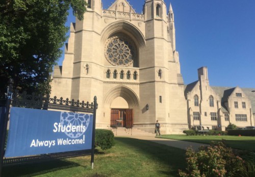 A large stone building with a rose window and a sign reading "Students Always Welcome!" in front.