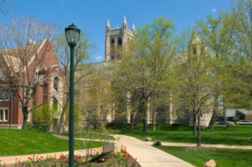 A sunny campus scene featuring a path, trees, and a historic building with a tall tower in the background.
