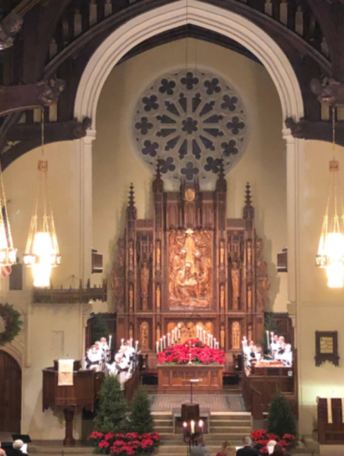 Interior of a church featuring a decorated altar, candles, and a large stained glass window, with choir members in white robes.