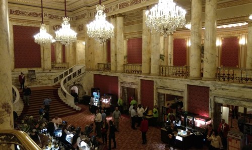 Elegant hotel lobby with chandeliers, marble columns, and guests mingling near display tables.