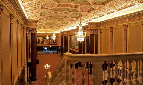 Elegant interior of a grand staircase with ornate ceiling, chandeliers, and decorative walls in a historic building.