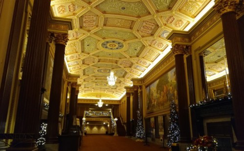 Elegant interior of a grand hall with ornate ceiling, chandeliers, and festive decorations, including Christmas trees.