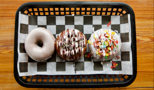 A basket with three donuts: a plain glazed, a chocolate-drizzled, and one topped with colorful cereal.