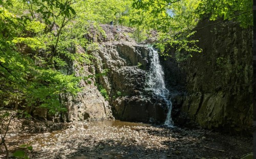 A serene waterfall cascades over rocky terrain, surrounded by lush green trees and a calm pool below.